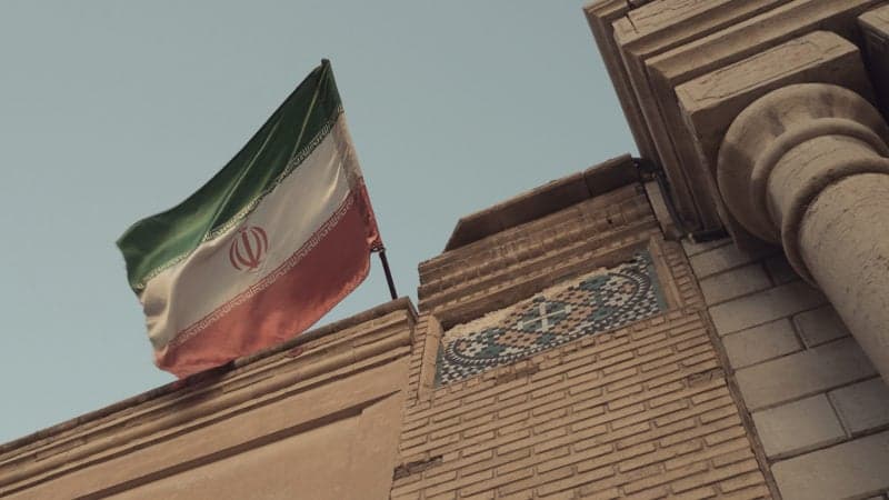 La bandera de Irán ondea sobre un edificio con arquitectura tradicional, contextualizando la noticia sobre la plataforma de criptomonedas iraní Nobitex y sus conexiones políticas.