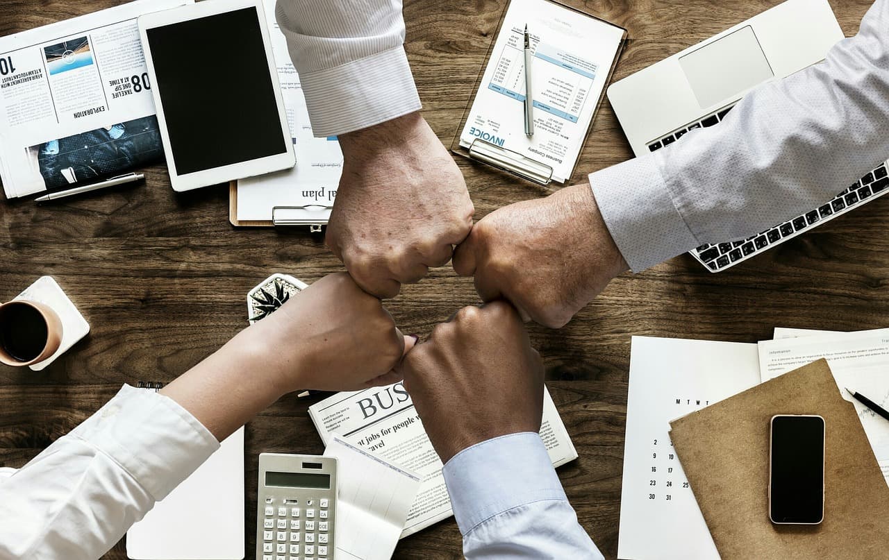 Vista cenital de un grupo de personas de negocios uniendo sus puños sobre una mesa de trabajo rodeada de documentos, dispositivos electrónicos y material de oficina.